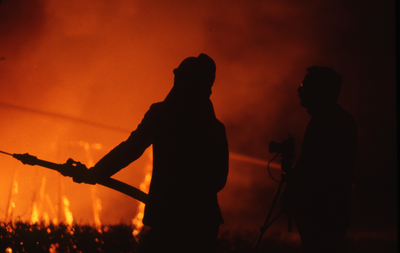 806138 Afbeelding van het silhouet van een brandweerman tijdens het blussen van de in brand staande houten gebouwen van ...
